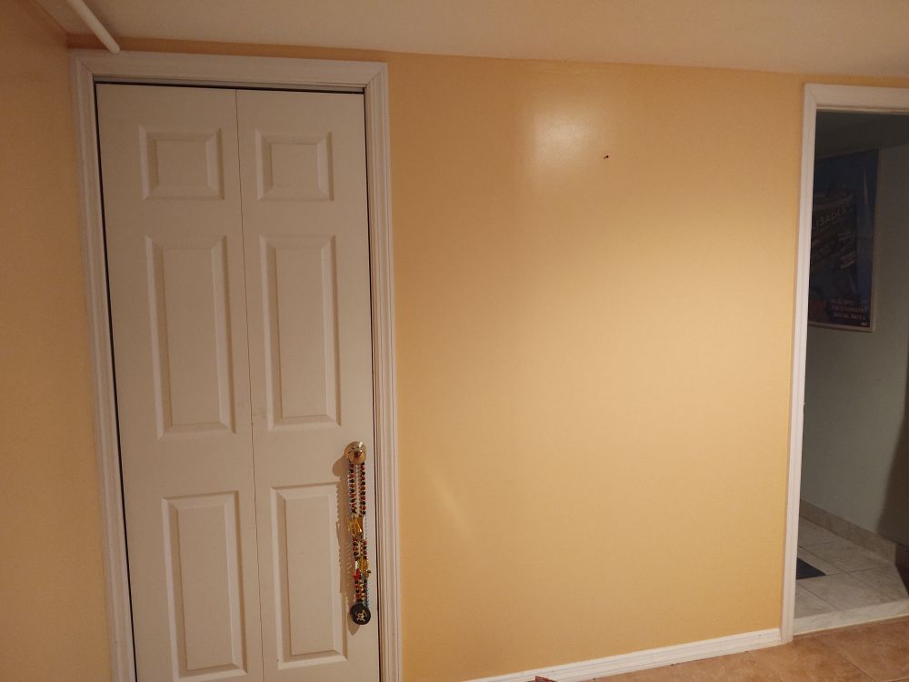 Wall of our basement freshly painted a warm peach tan color. You can see the open door to a bathroom on the right and a white door on the left. New Orleans beeads hang on doorknob on the left 
