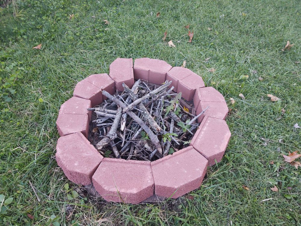 A irregular circle of red retaining wall bricks. One brick is roughly perpendicular to the others, giving the pit almost a spiral appearance. Dry branches are piled up inside it. 