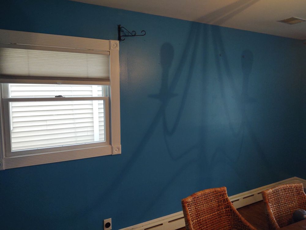 Wall of our dining room freshly painted a bold blue color. There is a window with blinds on the left. In the right front, you can see the tops of two wicker chairs. 