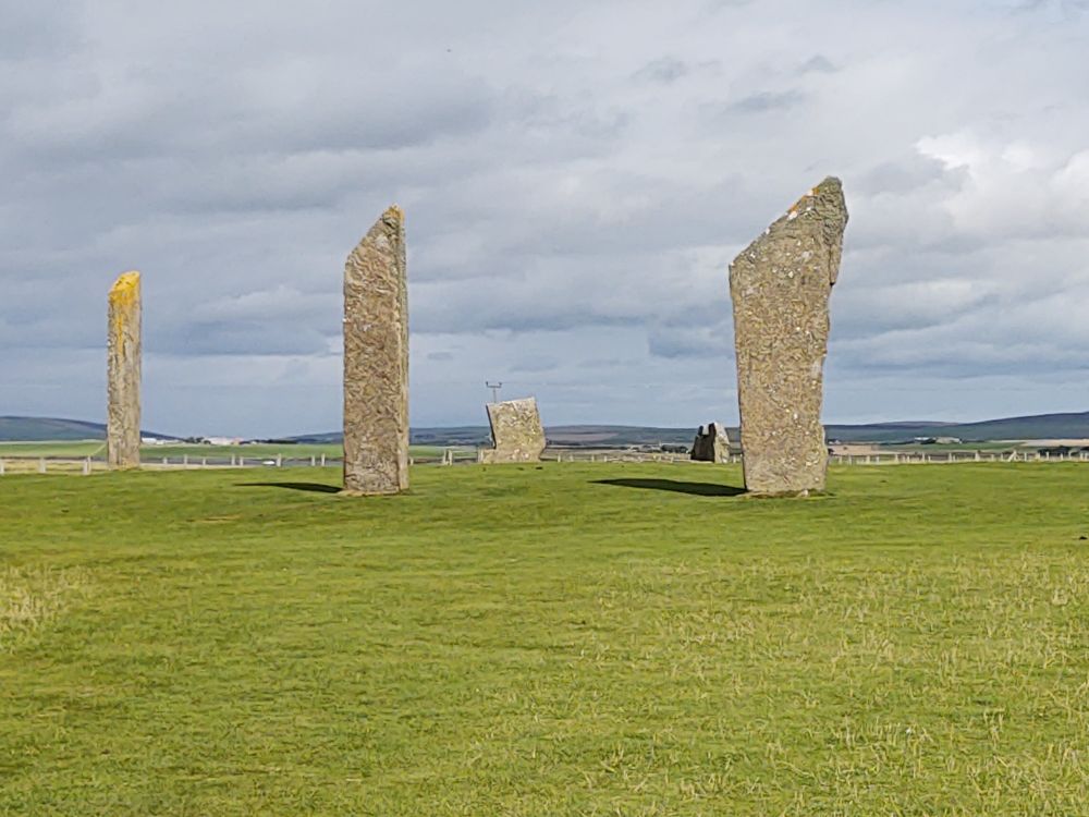 Several standing stones in a ring. These are the Stones of Stenness on Orkney. Three are tall and the tops slant to a triangular point. Two stones in the back are broken and much shorter than the other three. There is a field of short grass in front. The sky is gray with heavy clouds. 