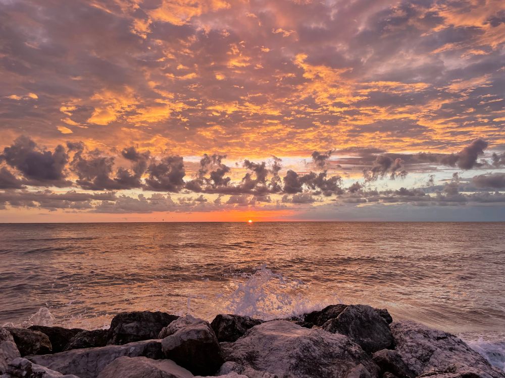 Das Bild zeigt eine stimmungsvolle Meereslandschaft bei Sonnenuntergang. Im Vordergrund sind dunkle, große Felsen am Ufer zu sehen, gegen die Wellen schlagen und Gischt aufspritzt. Dahinter breitet sich das ruhige Meer bis zum Horizont aus. Am Horizont ist die Sonne gerade dabei, im Meer zu versinken – sie leuchtet in einem kräftigen Orange. Der Himmel ist von zahlreichen Wolken bedeckt, die in warmen Farbtönen von Orange, Rosa, Violett und Gold angestrahlt werden und ein eindrucksvolles Farbspiel erzeugen. Die gesamte Szene wirkt friedlich und gleichzeitig kraftvoll.