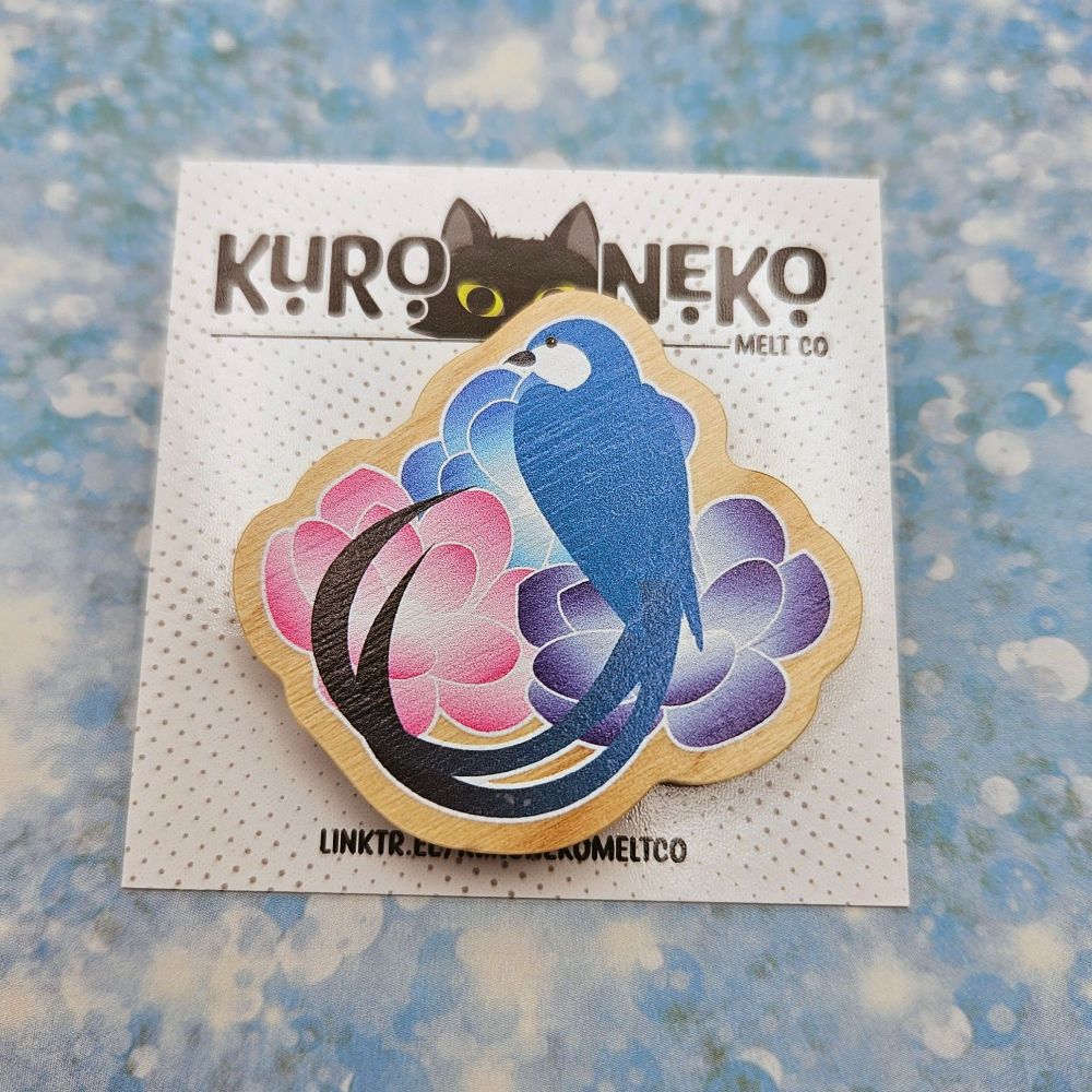 A wooden pin in a vector style of Meteion from Final Fantasy XIV. 

Meteion is a blue bird, sitting on three flowers - one blue, one purple, one pink. She has long tailfeathers that fade into black at the tips.