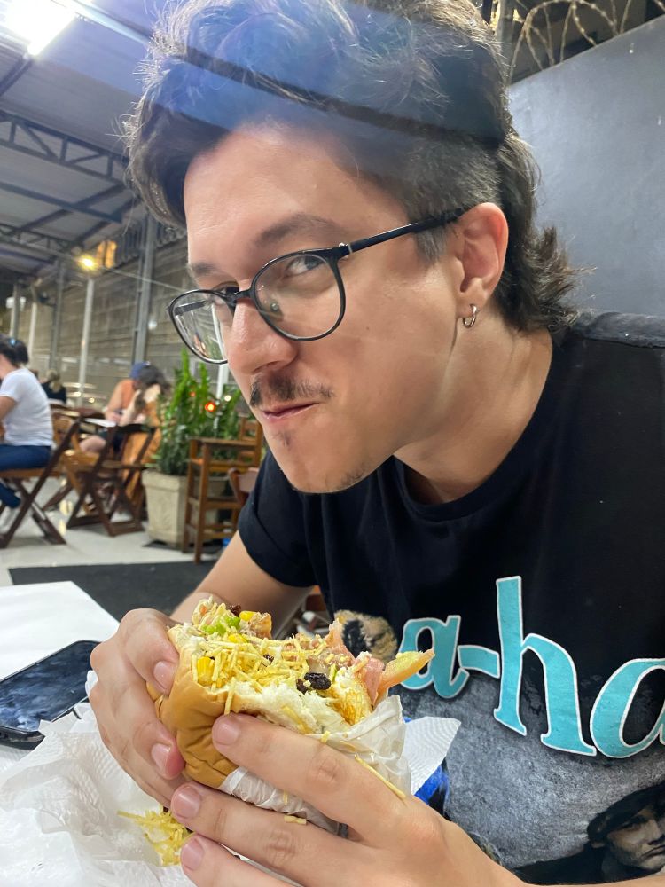 foto do pororocababy, meu namorado, segurando um hambúrguer enorme conhecido como pesadelo, usando uma camiseta preta com estampa do a-ha escrito em azul claro e parte de uma foto da banda aparece. ele está numa lanchonete com cadeiras e mesas de madeira e algumas pessoas desfocadas aparecem no fundo.