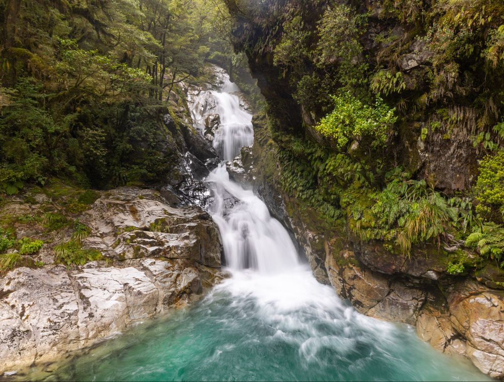 A waterfall cascading down a small canyon into a pool of clear blue-green water, bordered by granite and lush foliage.
