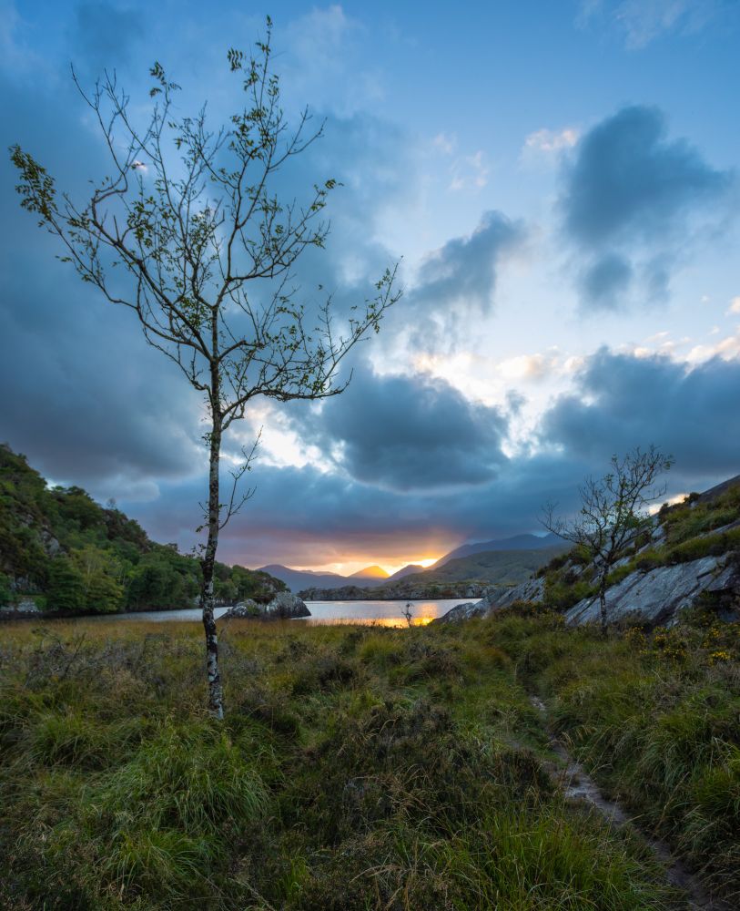 Sunset picture of a small lake on Moll's Head Drive in Ireland, with moody clouds and the sun going down behind the hills.