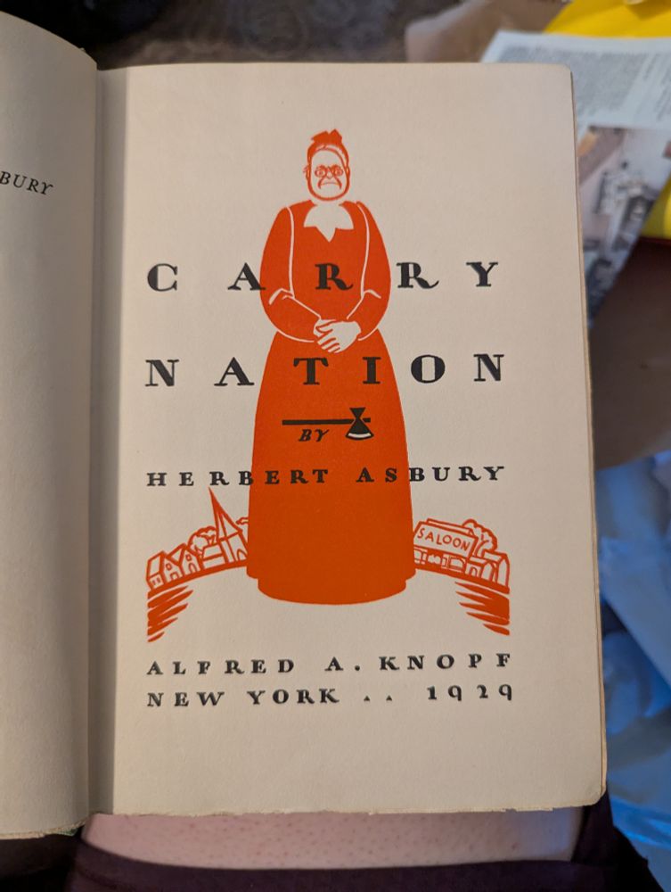 Woodcut-like illustration in red of a frowning, severe dee woman with foldes hands standing in front of a town with a church on one side and a saloon on the other. The text reads "Carry Nation by Herbert Asbury, Alfred A. Knopf New York 1929"