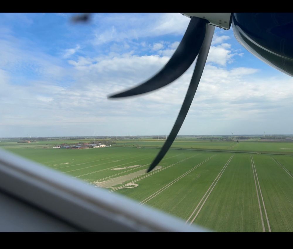 Foto aus einem kleinen Flugzeug raus beim Start. Man ist schon so 50m hoch. Sieht im Hintergrund die Felder und im Vordergrund Teile des Fenster inkl. Rahmen sowie den sich drehenden Propeller. Das ist der kürzeste Linienflug Deutschlands.