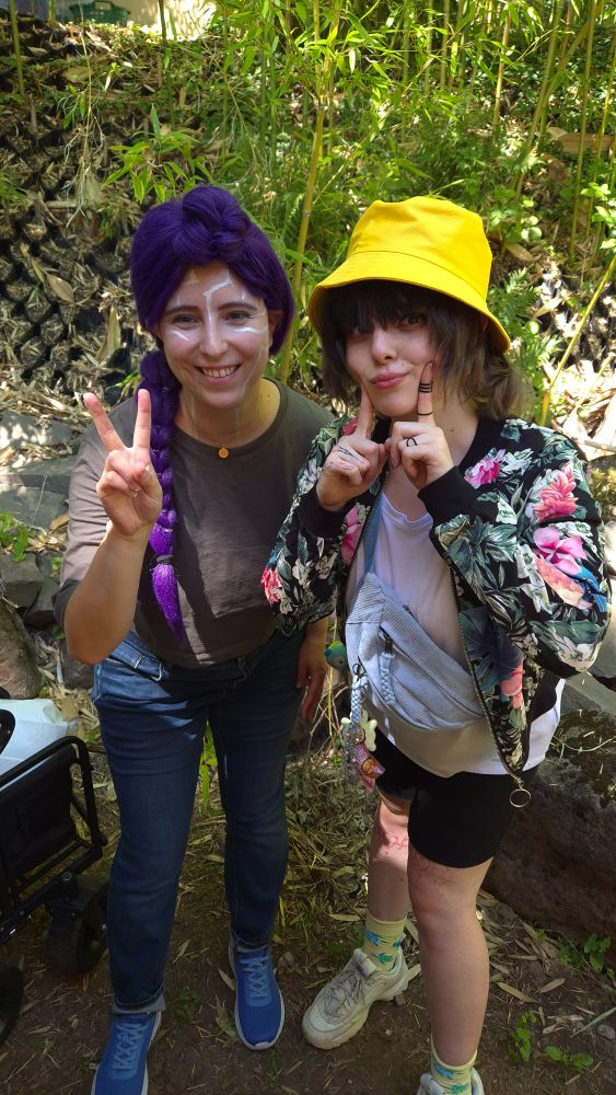 Photo of me as Rumi in her crop-top chill outfit and another cosplayer as Zoey in her undercover outfit, posing for the camera in front of some greenery.