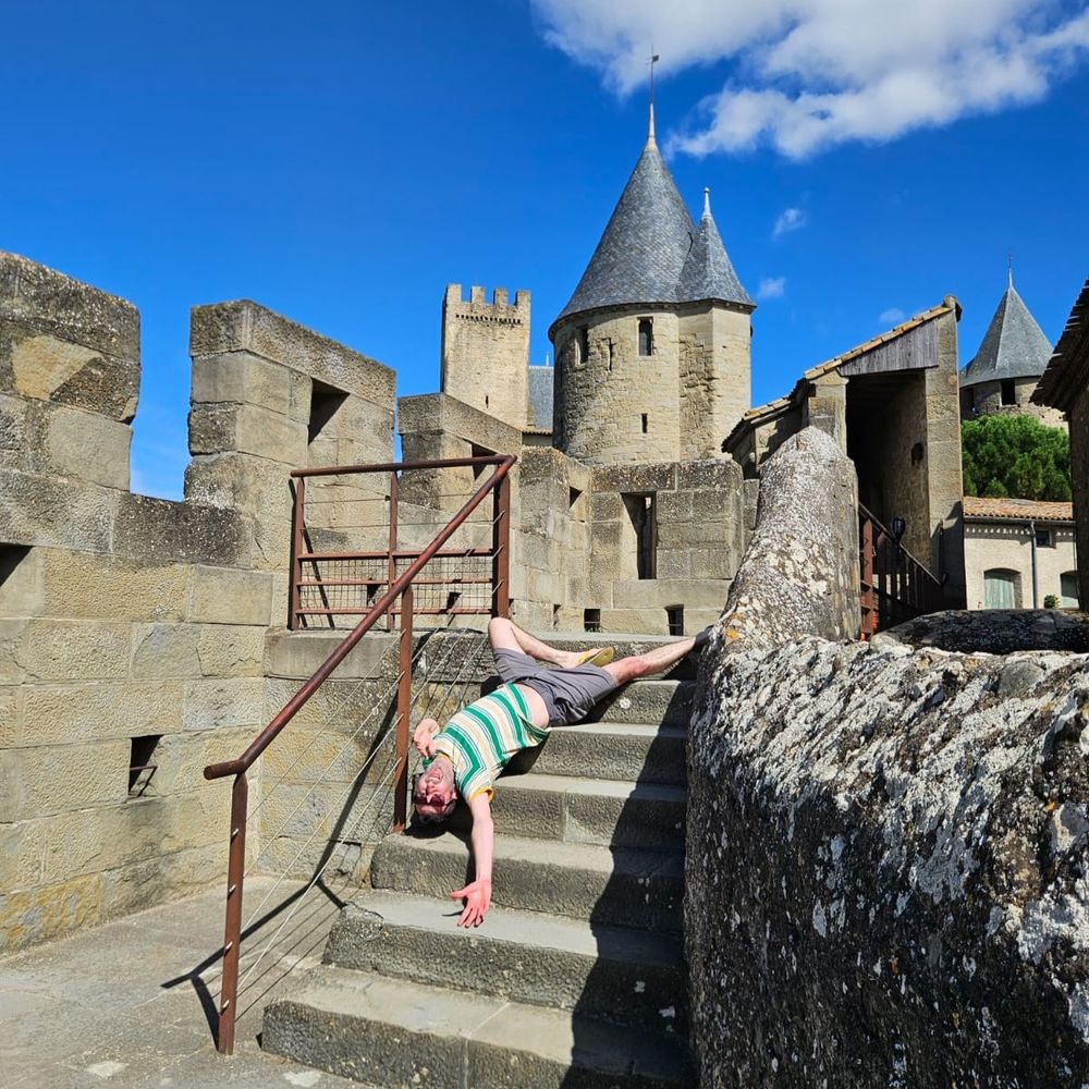 A man in a striped green and white T-shirt pretends to fall down a short flight of stone stairs. In the background are turrets 