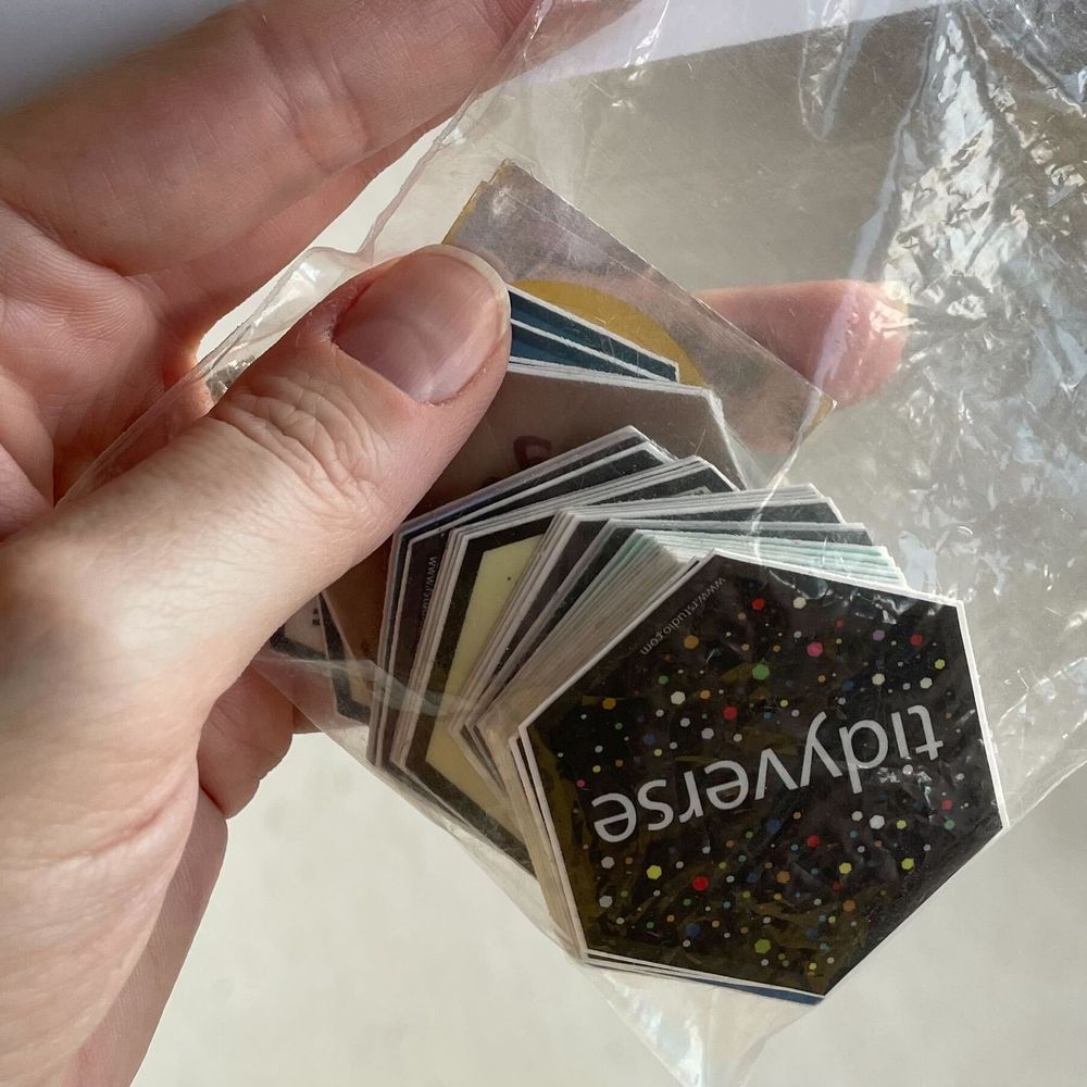 A left hand holds a small plastic bag full of small hexagonal "hex" stickers. The sticker visible on the top is a black sticker that says "tidyverse" in white letters and is covered in multicolored dots.
