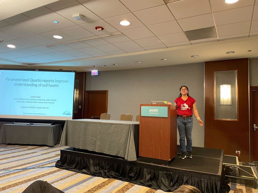 An image of Jadey Ryan presenting her talk at Posit conference 2023 in Chicago. Jadey wears a red t-shirt, jeans, and tennis shoes. She stands at a podium and smiles. On a screen beside her, you can see a slide saying "Parameterized Quarto reports improve understanding of soil health"
