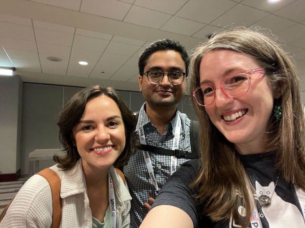 Three people pose, smiling, for a selfie. Natalia is on the left, a woman with short brown hair. She's smiling and wearing a cream colored sweater with a white Posit lanyard around her neck. Sam stands in the center. He's a man with black hair, a black and white checkered button-up shirt, a backpack, and a Posit lanyard. Libby is on the right, a smiling woman with brown and grey hair. She's wearing pink glasses, a dark grey t-shirt with a white cat on it, and a silver necklace.