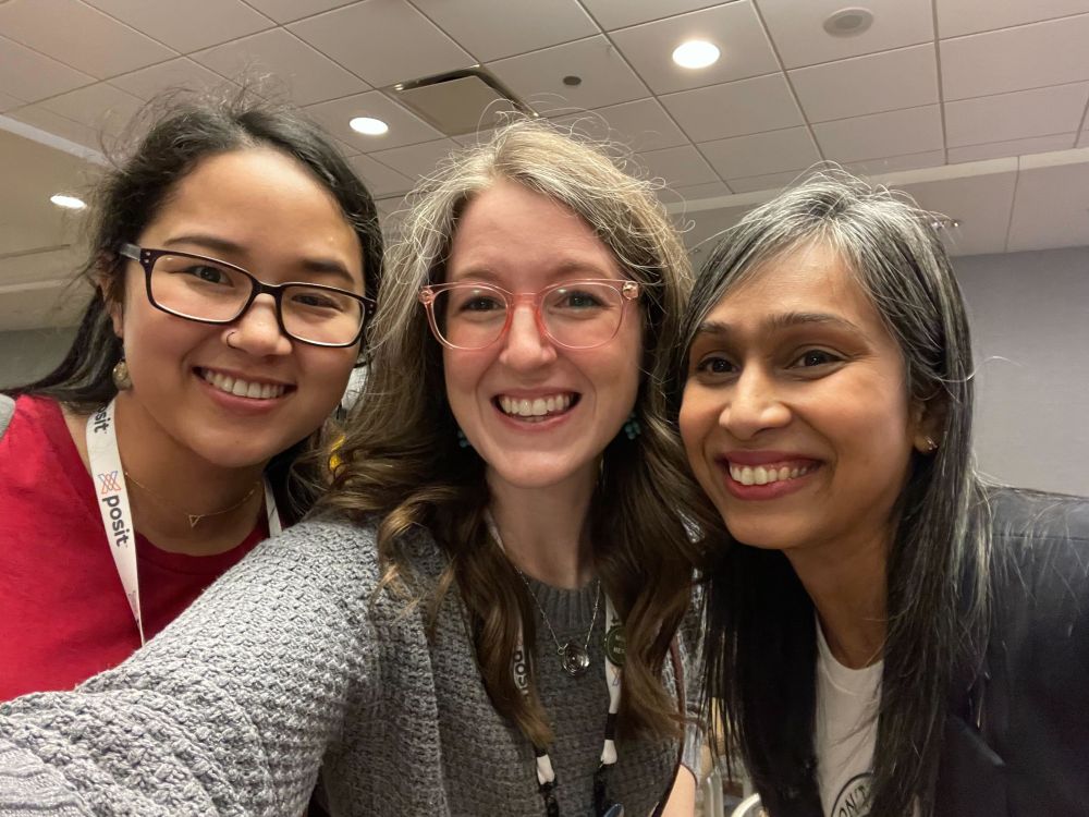 Three women smiling together for a selfie. Jadey on the left has black hair, dark rimmed glasses, a red t-shirt, and a white Posit lanyard. Libby in the center has brown and grey hair, pink glasses, and a grey sweater with a silver necklace. Deepsha on the right has black and grey hair, a t-shirt, and a blazer over it.