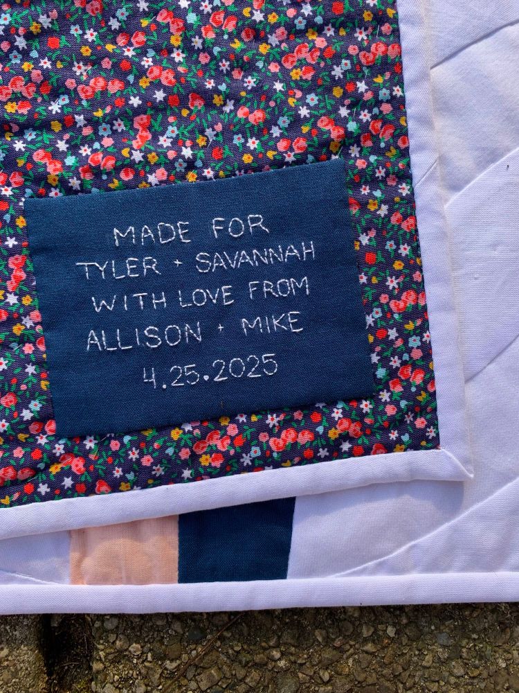 The back corner of the quilt with the quilt label; part of the front of the quilt can be seen underneath. The back is a colorful, small floral print. The label is navy, with following inscription stitched in white: “Made for Tyler + Savannah with love from Allison + Mike 4.25.2025”