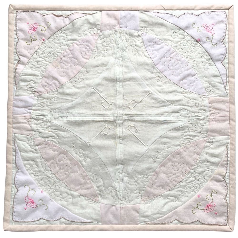 A double wedding ring quilt square in shades of ivory and pale pink. The outside corners have small pink flowers with dainty green stems. The center has four “J”s embroidered in white. The quilted stitches are in the circular, floral design from the tablecloth used for the backing of the piece.