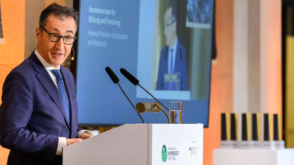Ein Mann mit schwarzen Haaren, Brille und einem blauen Anzug (Cem Özdemir) steht hinter einem Redepult und spricht. Im Hintergrund kann man auf einer Leinwand „Bundesminister für Bildung und Forschung“ auf Deutsch und Englisch lesen.