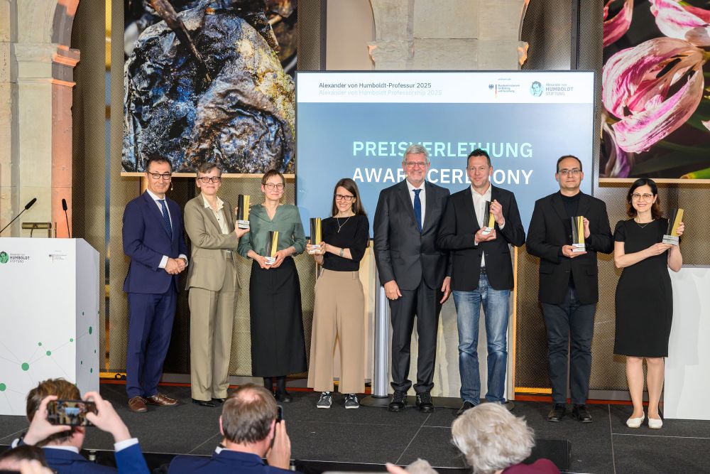 Ein Gruppenbild mit acht Personen: Cem Özdemir, Robert Schlögl und die sechs Preisträgerinnen und Preisträger mit ihren Stelen stehen auf einer Bühne und lächeln. Im Vordergrund sieht man Personen, zwei davon machen ein Foto mit ihrem Smartphone
