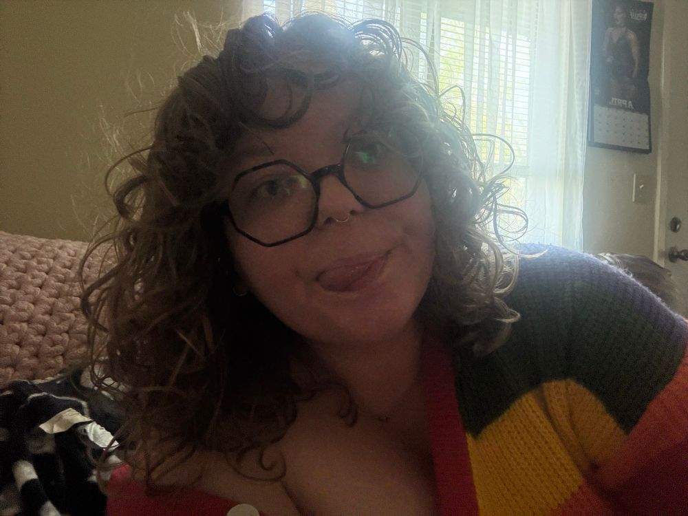 a photo of me. my hair is laying on my shoulder, dirty blonde/light brown & curly. i have bangs! i'm wearing brown hexagon glasses and i have my tongue sticking out the side of my mouth. i have a gold circular septum piercing and im wearing a rainbow sweater! the background is a beige wall with a window that has sheer white curtains. next to the window is a wwe calendar😂