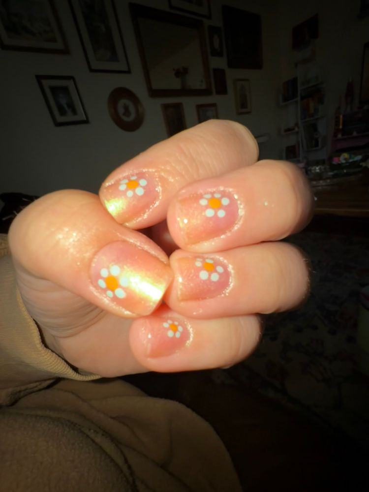 .5 photo of my left hand nails. they are a light skin toned esq pink with a green iridescent shift. there is nail art in the corner of each nail of a blue dotted flower with a yellow center 