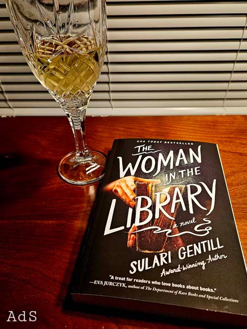 Book: The Woman in the Library by Sulari Gentill next to a glass of white wine/AdS