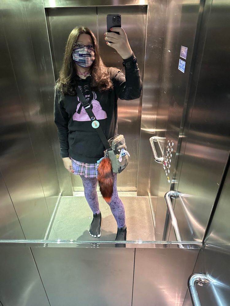 Selfie of me wearing a black drone “beep” hoodie, pink collar, Elizabeth Line face mask, purple plaid skirt, purple patterned leggings, and black toe shoes