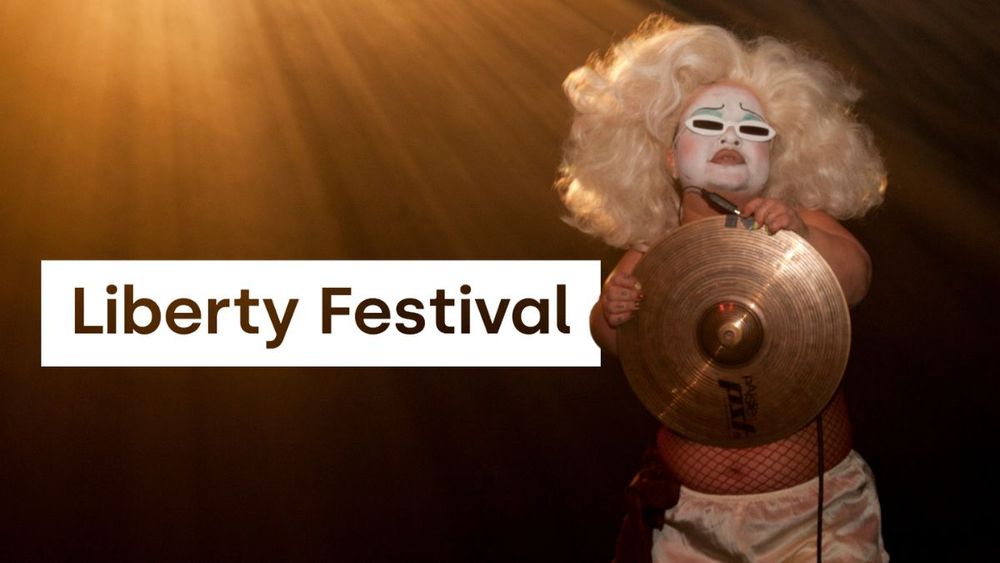 Liberty logo is digitally imposed behind a production shot. Midgitte Bardot, a person with dwarfism, performs on stage. They wear bold white makeup with blue eyeshadow covered mostly by thin white sunglasses, a glamorous blonde wig, red fish net tights and silky white shorts. They hold a cymbal in front of their chest which reflects golden light, with rays of more golden light flooding from the top left. Photo by Holly Revell. 