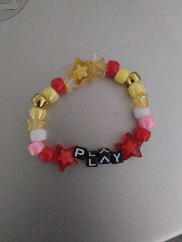 a kandi bead bracelet reading "PLAY" with the a being an upside down letter v for stylization. the word is surrounded by red stars that go into single red, pink, and white beads, a smaller yellow star, and single gold, yellow and red beads. then there are two small yellow stars and one yellow star in the middle joining the two sides. the design is symmetrical.