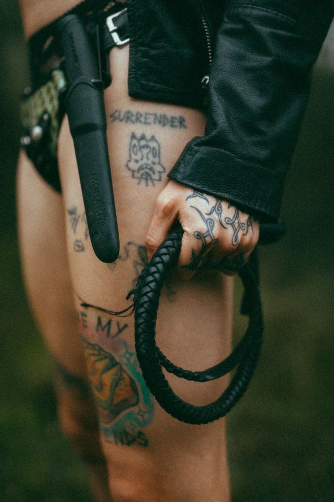 a similar shot to the one before except Charlotte is holding a small bullwhip encircled in her hand. her stick and poked tattoo reading "surrender" is prominently visible. 
