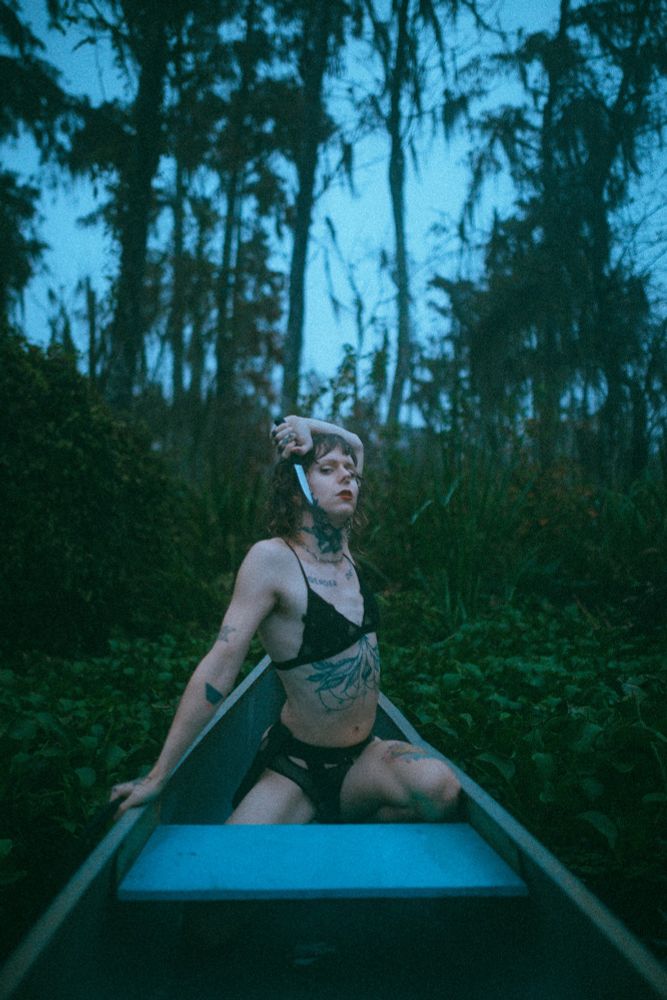 Charlotte poses at the bow of a canoe in the Louisiana swamp. She is holding a small hunting knife to her smoldering face wearing a black bra and panties. The photo is moody and blue toned, as was the weather that day. 