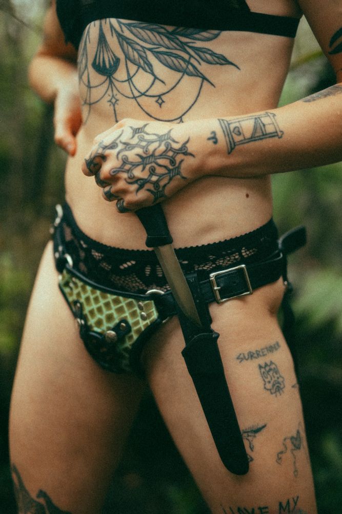 a shot of Charlotte unsheathing a small knife from a holster connected to her green alligator print strap on harness. you can see her menagerie of tattoos across her hands, thighs, and ribs.