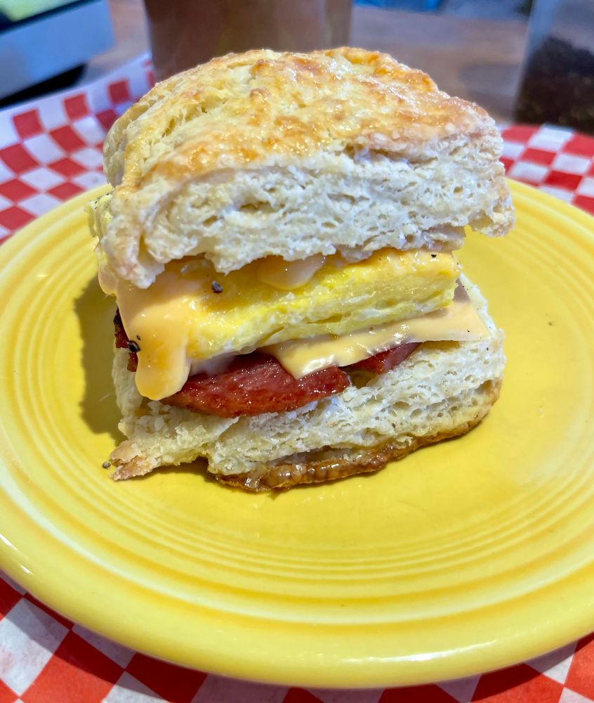 a close up of the pork roll egg and cheese biscuit sandwich. you can see the flakes of the biscuit, the crisp on the taylor ham, and molten cheese dripping from the rolled omelette. 