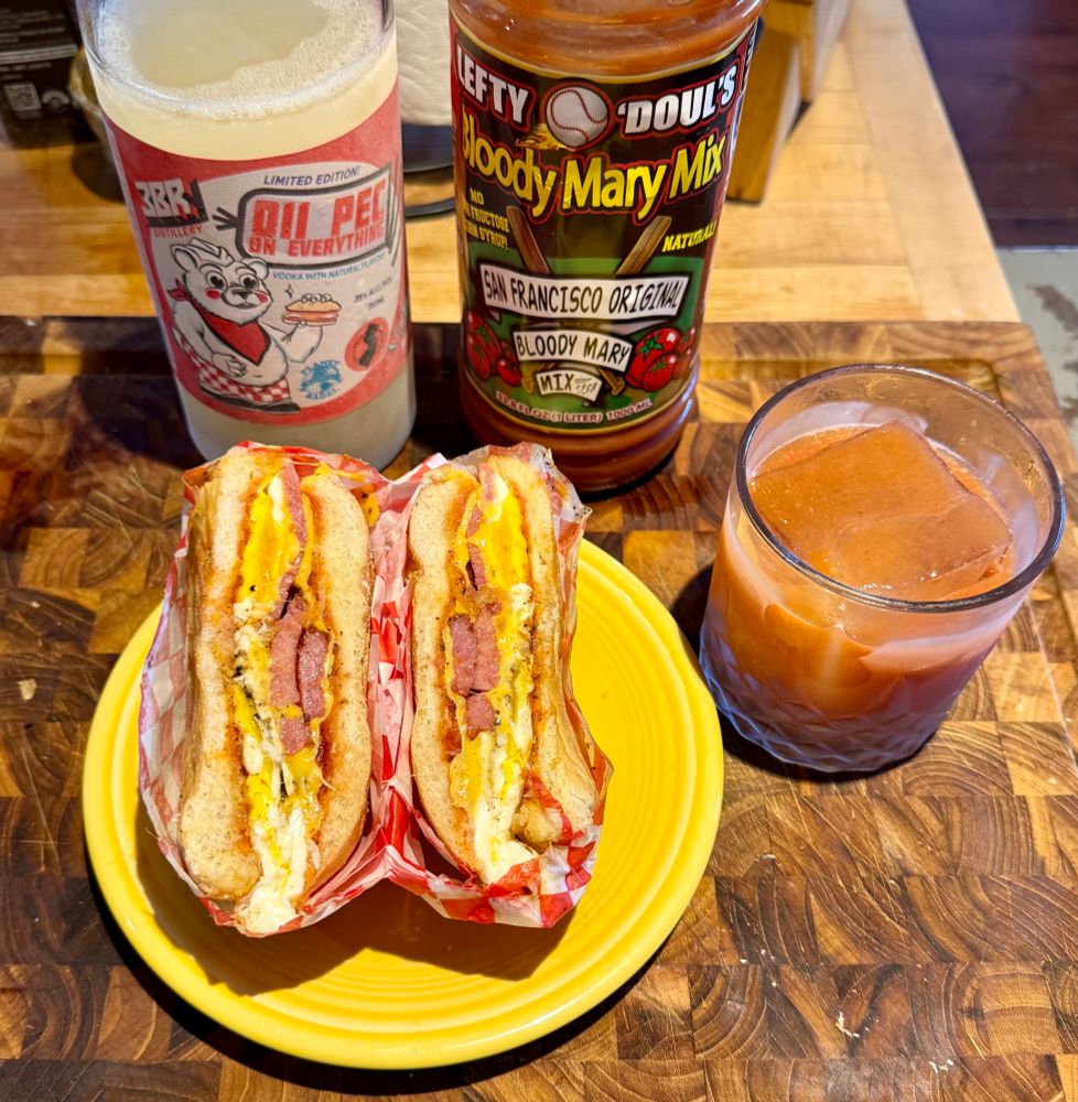 split porkroll egg and cheese on a kaiser roll showing the cross section, it’s wrapped in red and white checkered paper and sat on a yellow fiestaware plate. behind it is a frosty bottle of 3br 011 pec on everything vodka and lefty o’douls bloody mary mix; next to it is a shaken bloody mary in a frosty glass with one big cube. 