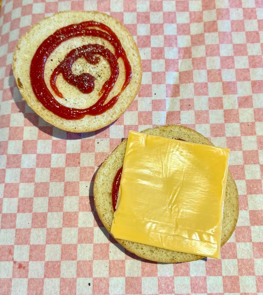 halved toasted kaiser roll, there’s a spiral of ketchup on both halves with salt and pepper, a slice of american cheese on one half