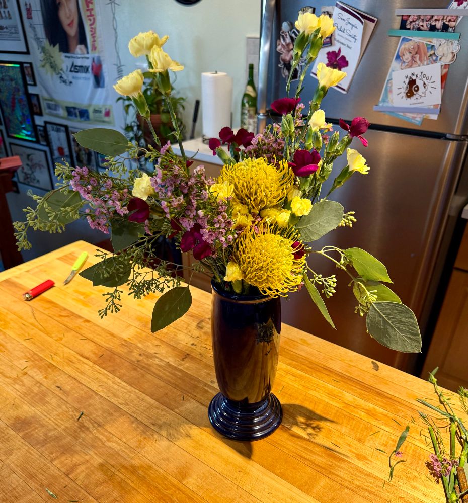 high angle of a flower arrangement in a blue vase. it has cream and dark red colored small rose looking flowers, and big yellow stringy orb looking flowers. there’s purple flowers, those are from the flowering rosemary sprigs. there’s also some big leafy greens poking out. the big yellow orb flowers are arranged in the middle, the rest is blowing out to the sides above them. 