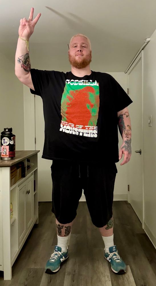 bathroom selfie; i’m wearing black shorts, white socks, and green new balance sneakers. i have a black shirt on with a green and red heat signature image of godzilla, it says godzilla above it and king of the monsters below it. 