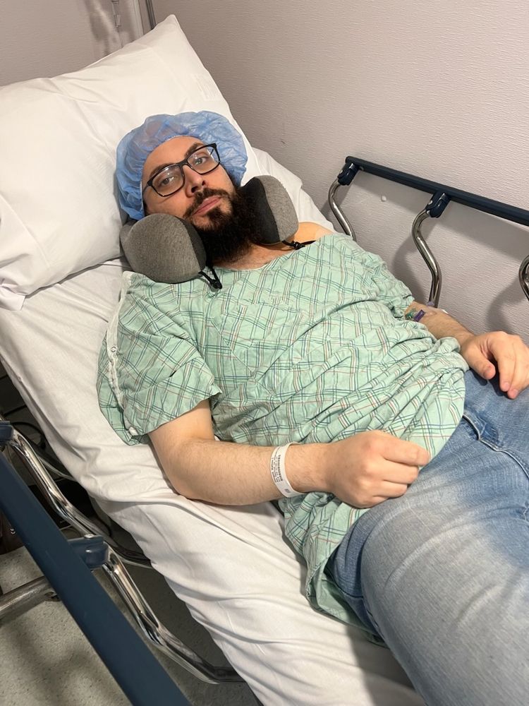 A bearded man lying in a hospital stretcher with a green hospital smock instead of a shirt and a blue hairnet unevenly placed over his head.