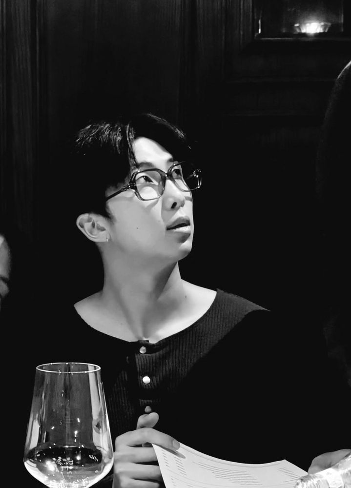 Namjoon in black wearing a pair of glasses, looking off to the side