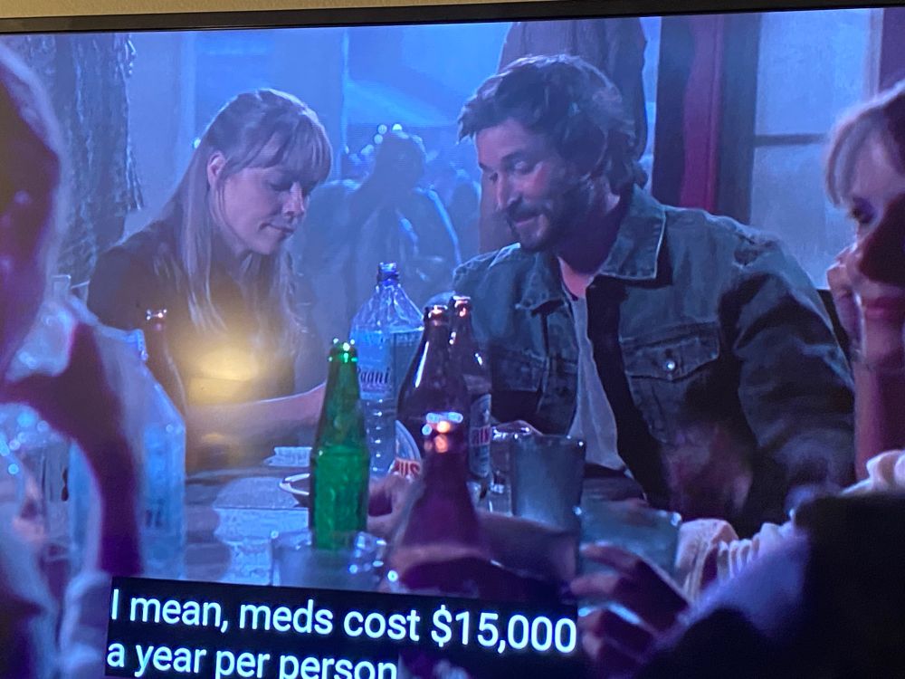 Dr. John Carter (Noah Wyle) sitting at a table with lots of beverages on it. Multiple people in the room including a blinde white woman he is next to.