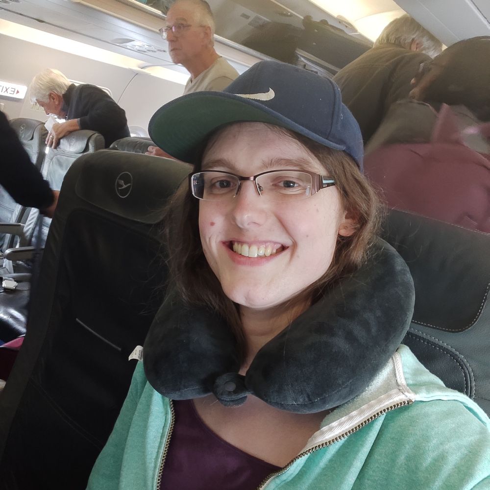 Kris, a woman with brown hair and rectangular glasses sitting in an aeroplane seat.

She is wearing a baseball cap and a neck pillow.