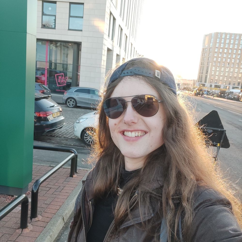 Kris, a woman with long brown hair, rectangular glasses, a backwards baseball cap, and a brown leather jacket, standing in a street with the sun behind her.

She is smiling.