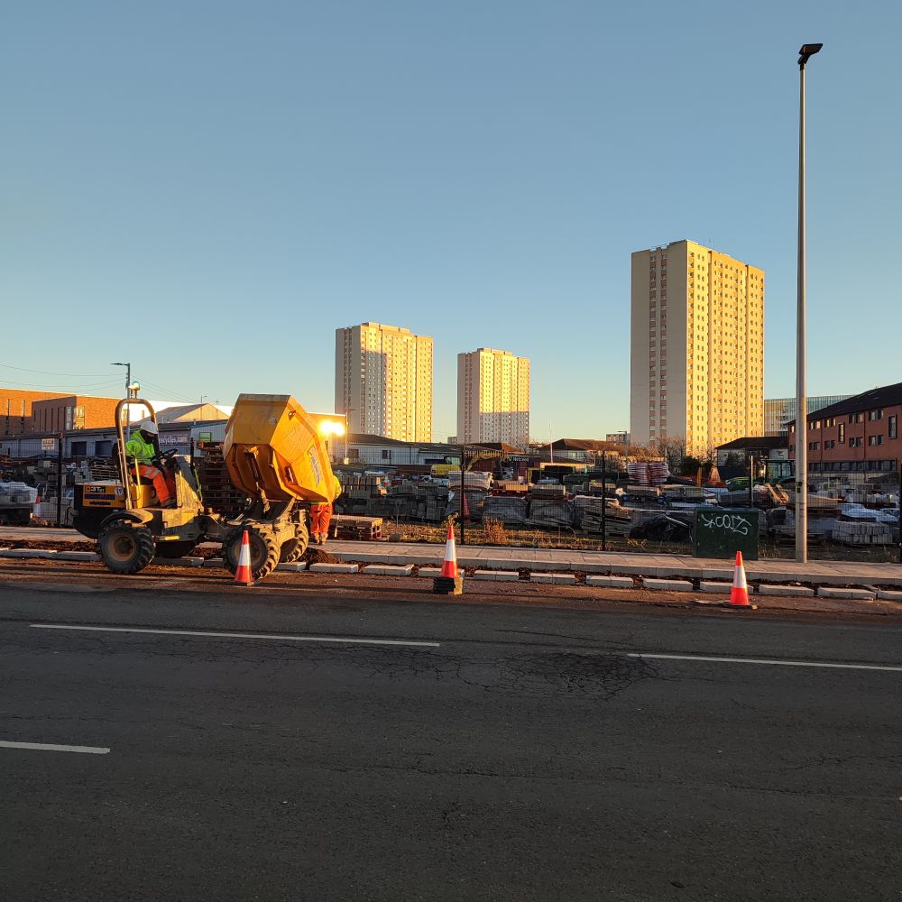 A photograph of 2 road maintenance workers and a small dump truck, working at the side of the road.

In the background are 3 tower blocks of flats.

It is dusk on a cold November afternoon in Glasgow.