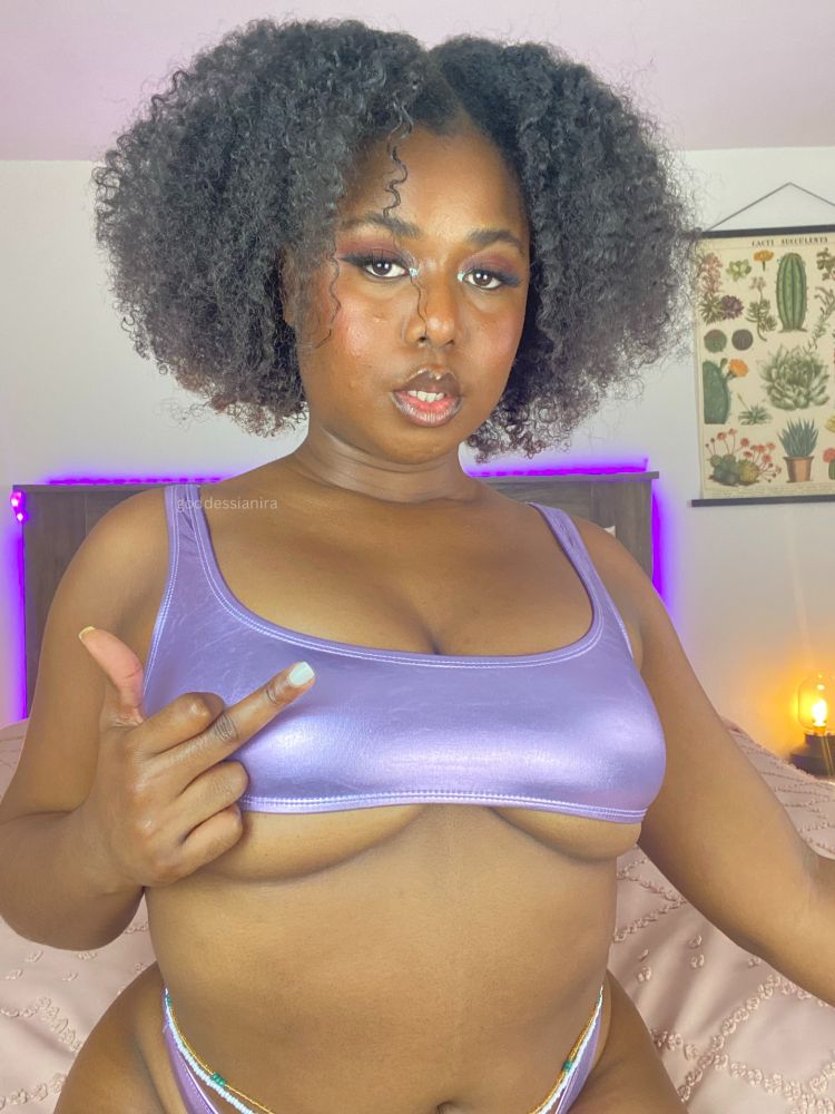 Goddess Ianira sits on Her bed in a shiny purple bikini. Her underboob and pretty tummy are on full display and you can't help but drool over them. She's wearing waist beads that draw your eyes down to Her curves. She's got Her middle finger resting on Her titty. Her nails manicured and painted white. She stares directly at you with a knowing look. She knows how weak you are for Her.