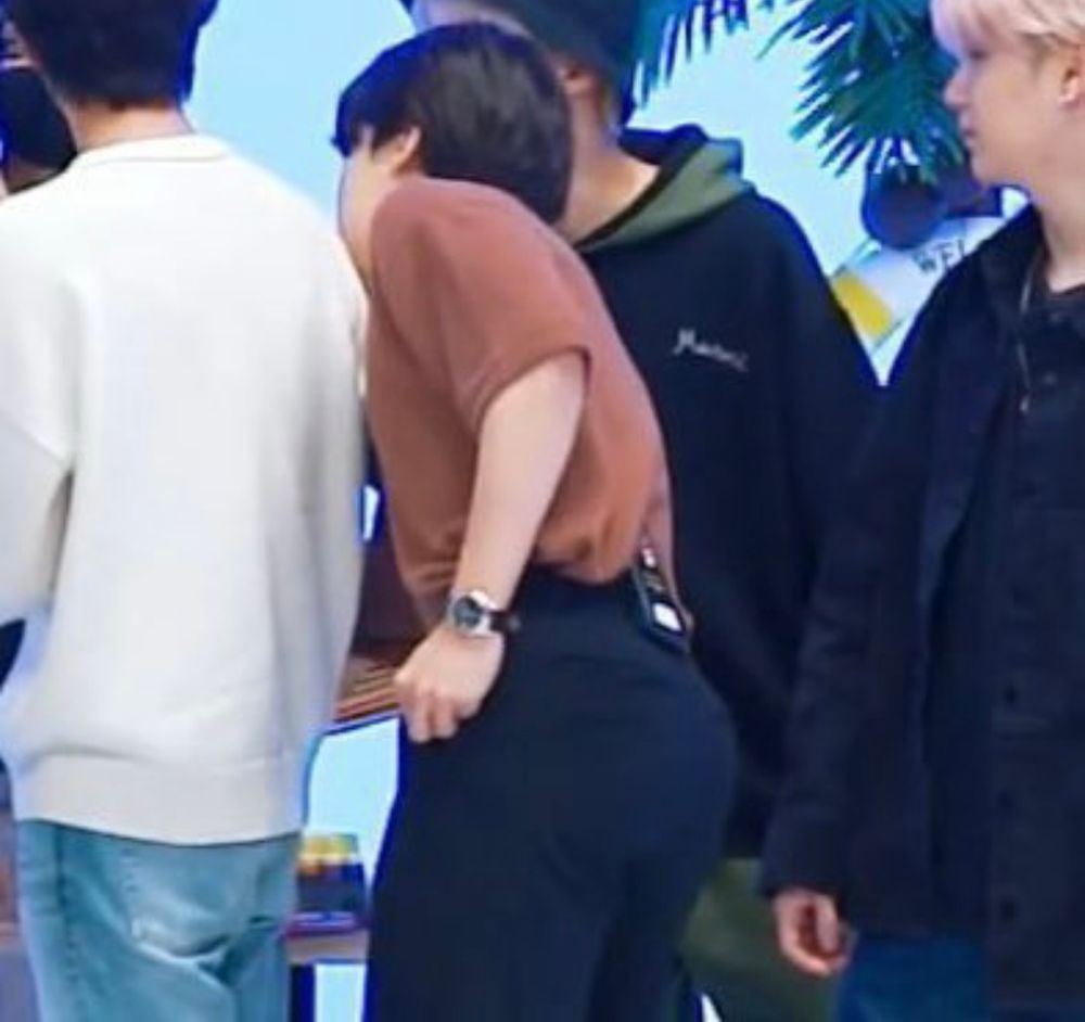 jimin facing away from the camera with an orange shirt and navy pants bending over slightly, his glorious ass on display.