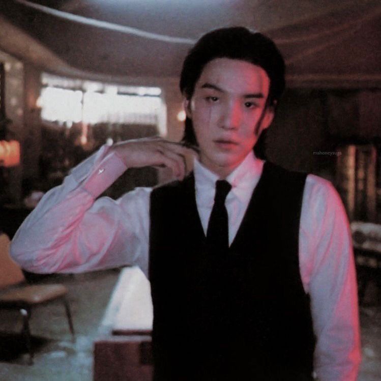 agust d in a white button down, black vest, and black tie. he tugs his collar as he gazes into the camera.