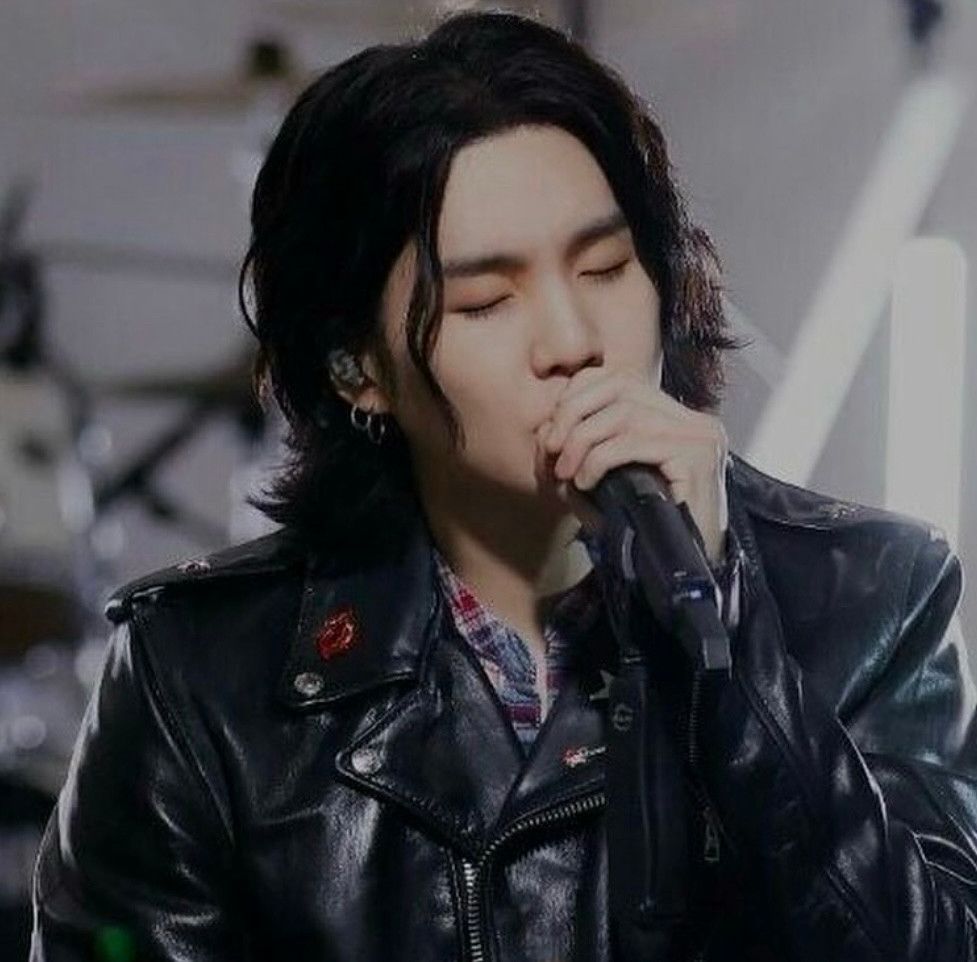 yoongi with a black leather jacket and long black hair. he holds onto a mic as he sings