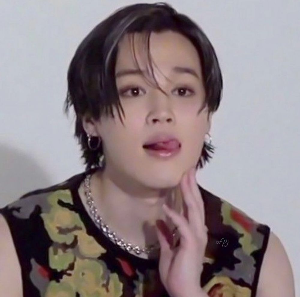 jimin in a black, yellow, red, and gray pattern sleeveless sweater. his hair is black and falls in his face. he has on a silver necklace and silver hoops. his tongue poking out.