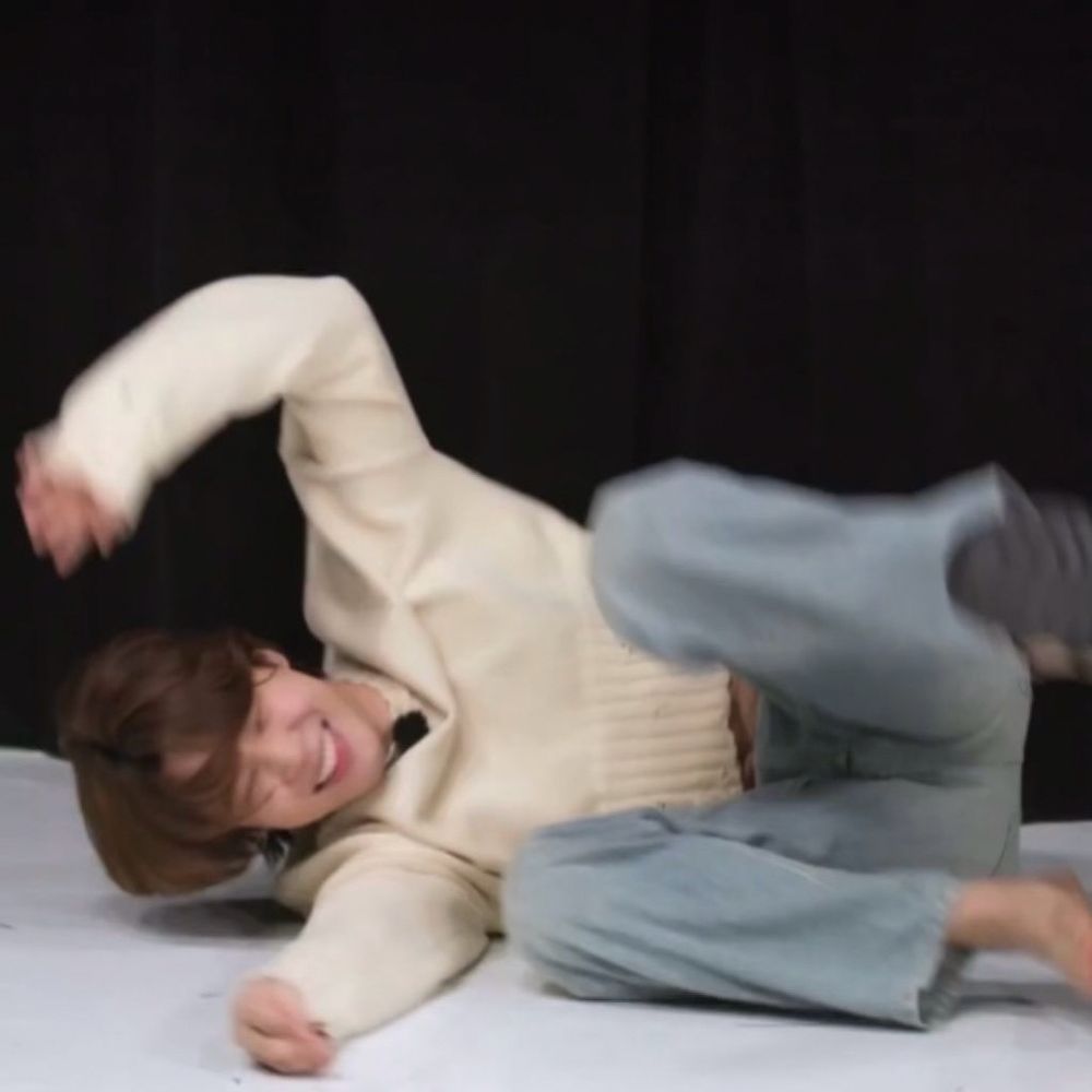jimin during an episode of run bts, mid falling on the floor. he has on a cropped cream sweater and light denim pants. he lost one shoe and his hair is long and brown.