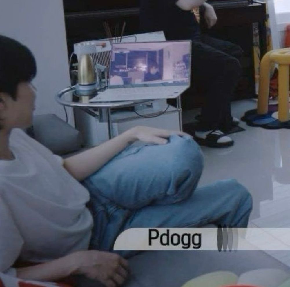 jimin during his production diary documentary sitting on pdogg’s couch with his knees up in blue jeans and a white shirt 