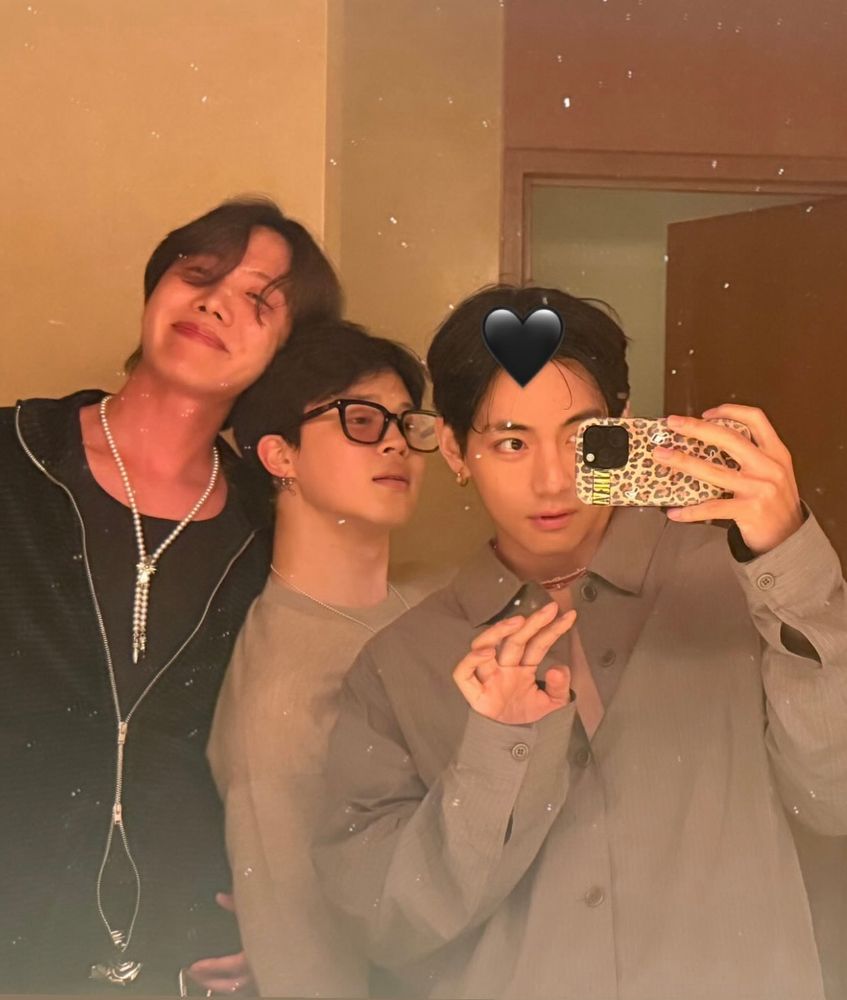 three absolute diva dolls taking a mirror selfie. hobi has on a black shirt and sweater with a pearl necklace, his long brown hair is windswept. jimin is leaning against him in a gray long sleeve shirt with short hair and glasses. taehyung takes the pic, his hair also short in a clay button up. he has his iconic cheetah print phone case. and a black heart over part of his hair.