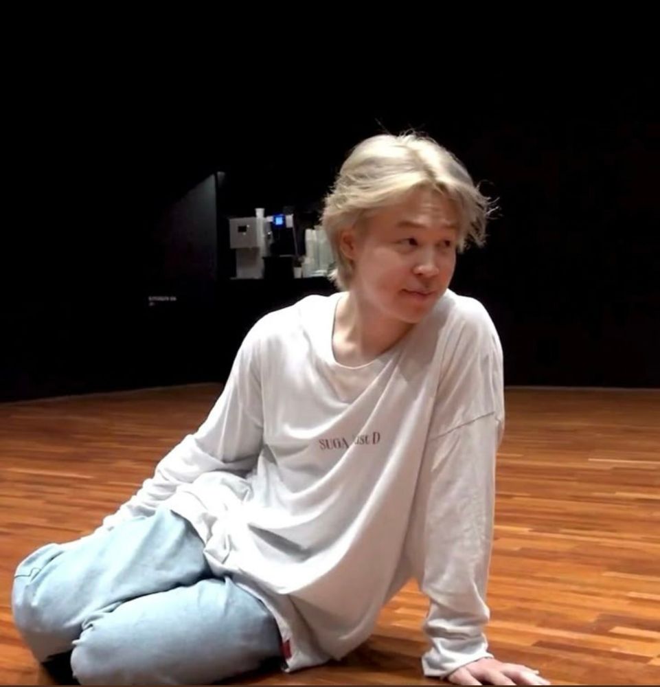 jimin sitting on the bts dance room floor in a white shirt, blue jeans and blond hair with his legs tucked together 